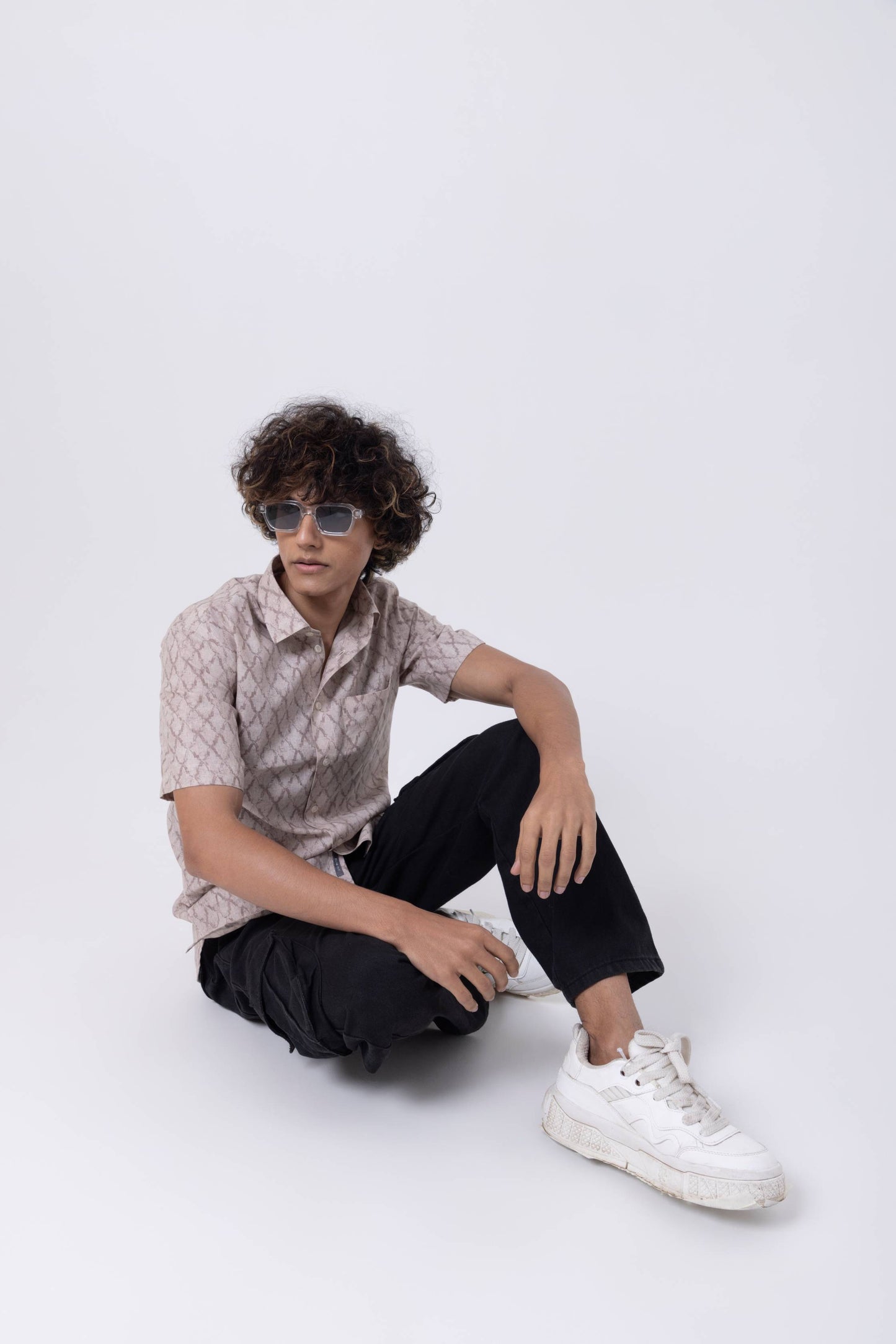 Stylish young man wearing a patterned shirt and sunglasses, sitting casually on the floor.